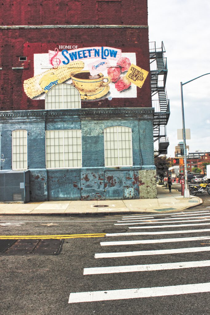 Bild im Hochformat von einer Straßenecke mit einem Haus im Fokus das aufmalte Werbung zeigt.Aus dem Artikel New York Reise 2019