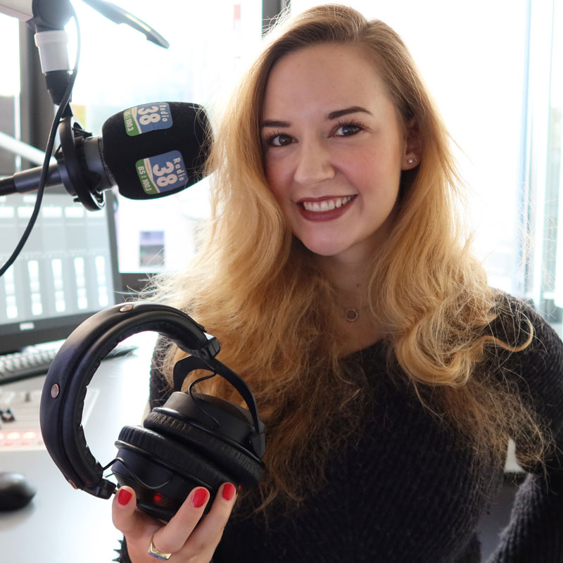Junge Frau sitzt in einem Radiosender vor einem Mikrofon und hält Kopfhörer in der Hand und lächelt in die Kamera. Aus dem Artikel: Wir fordern, dass der Frauentag 365 Tage im Jahr ist.