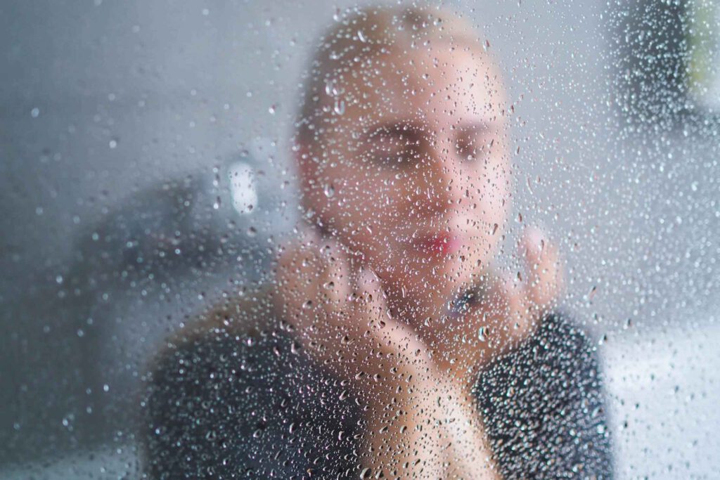 Junge blonde Frau hinter Duschwand mit Wassertropfen wischt sich Wasser aus dem Gesicht. Aus dem Artikel: Etwas ist jetzt anders. Panikattacke